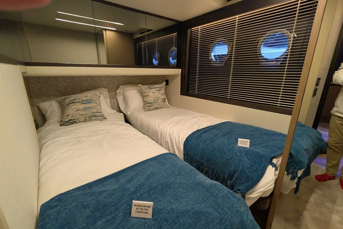Compact twin bedroom featuring two beds with blue throws, decorative cushions, and porthole windows; caution sign visible.